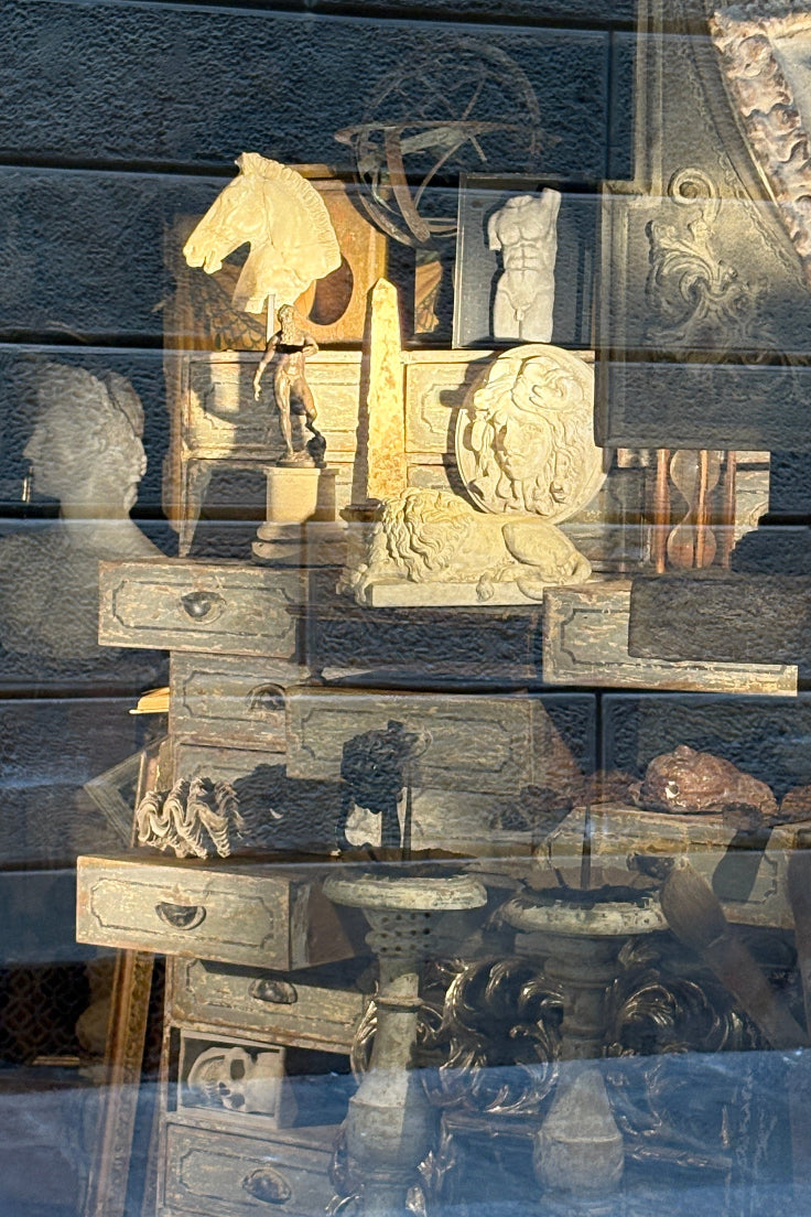 A display of assorted vintage and antique objects arranged on wooden stands behind a reflective window. Visible items include carved stone lion heads, a horse figurine, small drawers, and other decorative pieces, with strong outdoor light reflections across the glass.