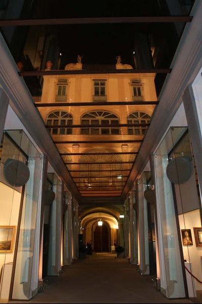 A long, symmetrical interior corridor with tall white columns, glass panels, and warm lighting, leading toward an arched opening in the background.