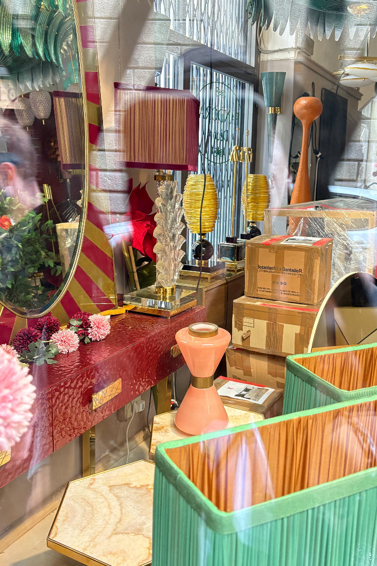 A busy retail display or showroom scene with multiple home decor items, including green fringed lamp shades, stacked cardboard boxes, a pink sculptural lamp, yellow table lamps, and a red textured console table.