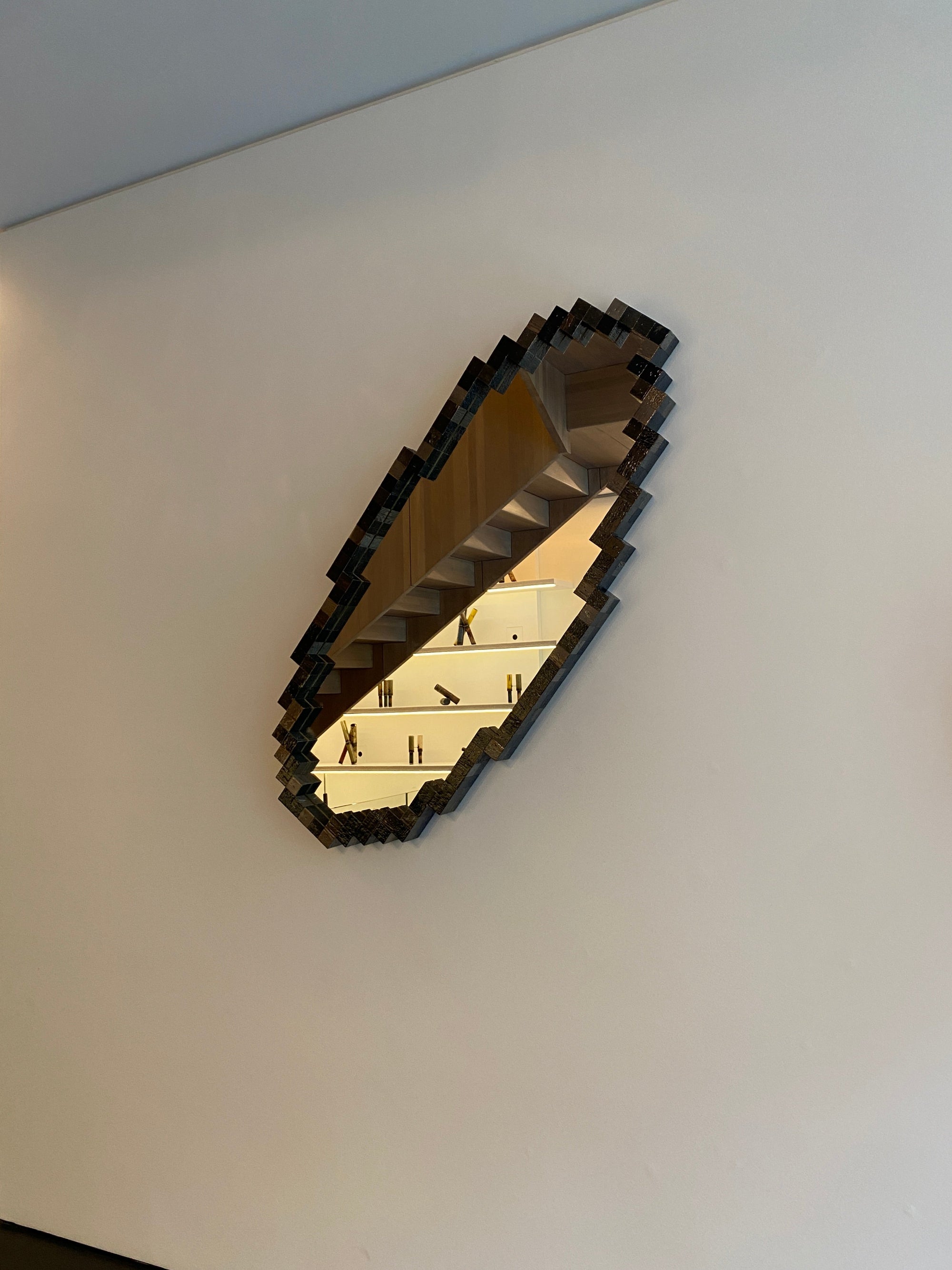 A modern, abstract wall mirror with a pixelated or blocky black outer frame and a reflective gold-toned interior, mounted on a plain white wall.