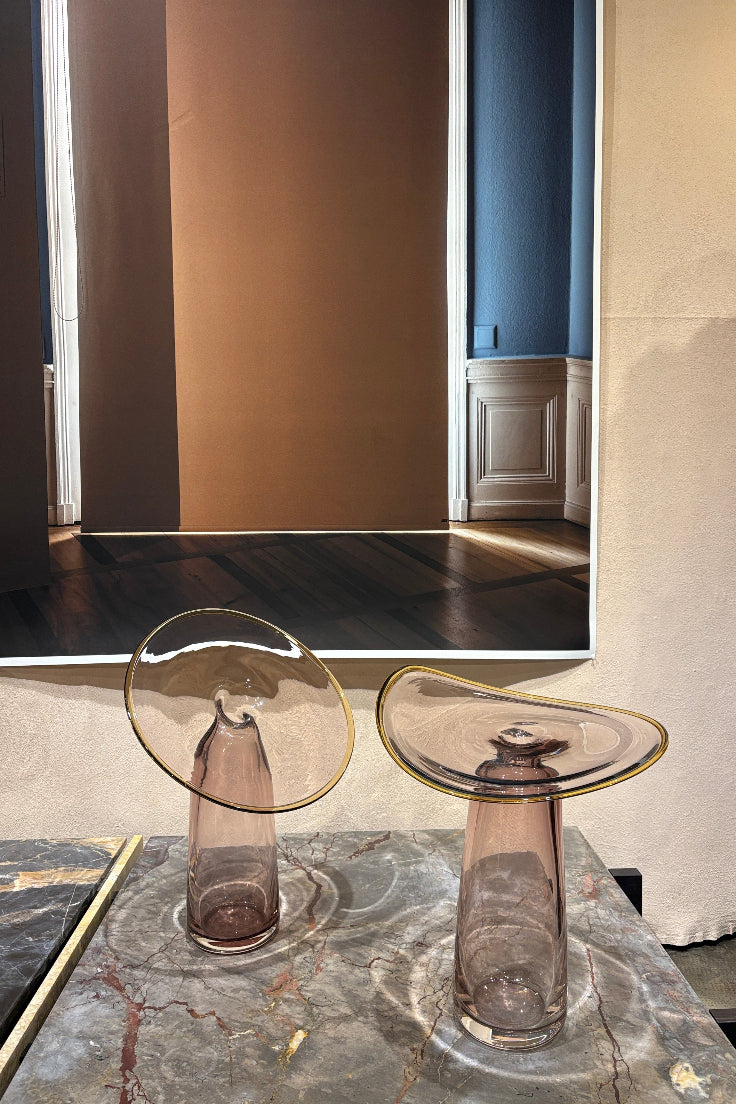 Two clear glass highball glasses with gold-rimmed edges and a smoky amber tint, shown on a dark marble countertop next to a mirror reflecting an interior space.