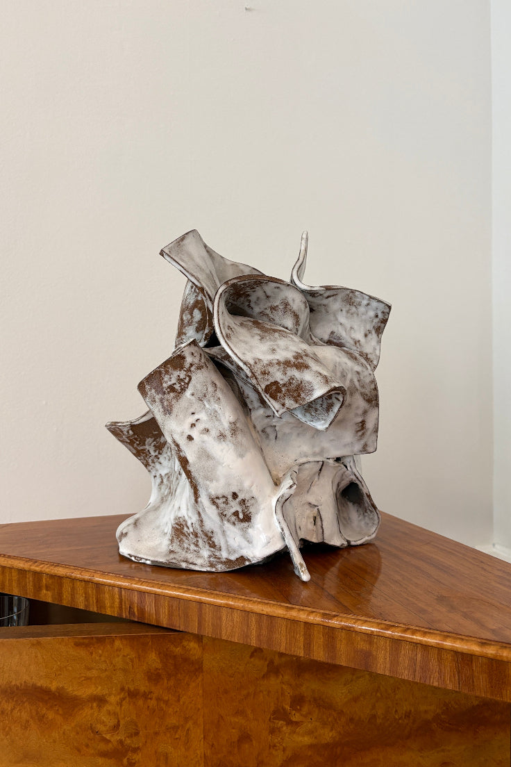 An abstract sculptural object with a mottled white and brown ceramic-like surface sits on a polished wooden table against a plain off-white wall.