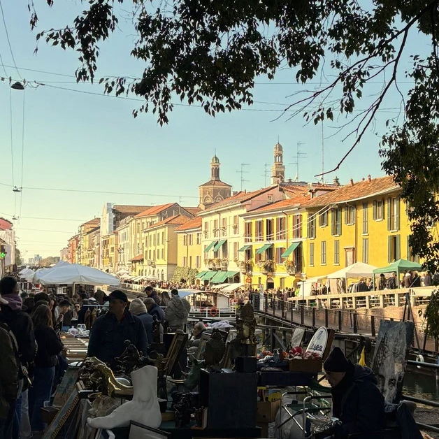 Outdoor market scene with people and stalls along a canal in an urban setting.