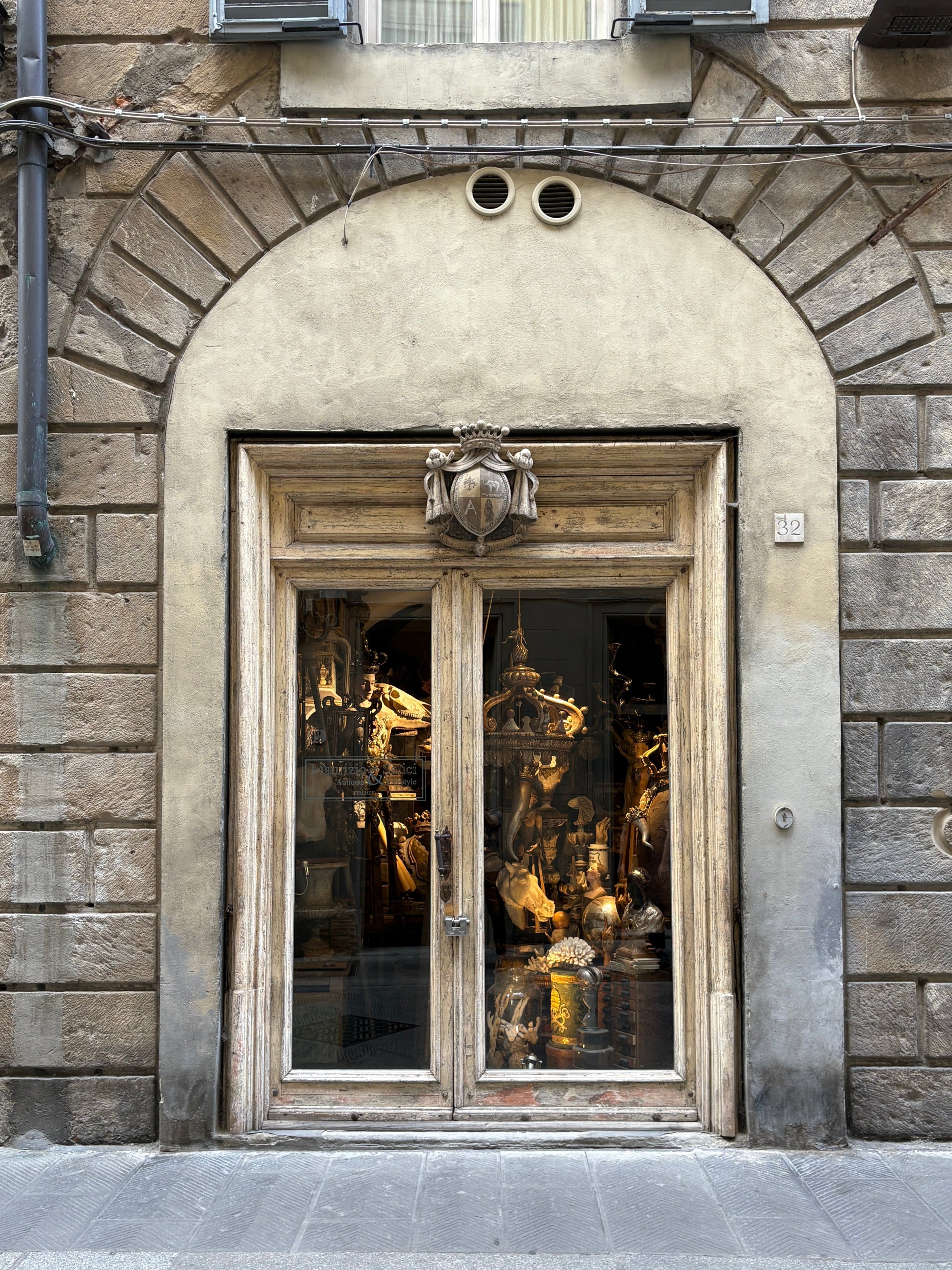An ornate stone building facade with a large arched doorway featuring double wooden doors and tall glass panes, reflecting warm golden light from an interior display.