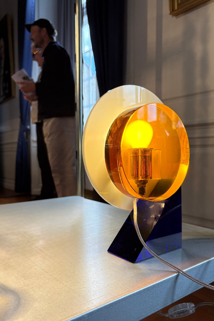 A modern table lamp with a glowing warm yellow bulb inside a translucent amber circular shade, mounted on a dark blue geometric base and set on a tabletop in an indoor room. A blurred person is visible in the background near a window.