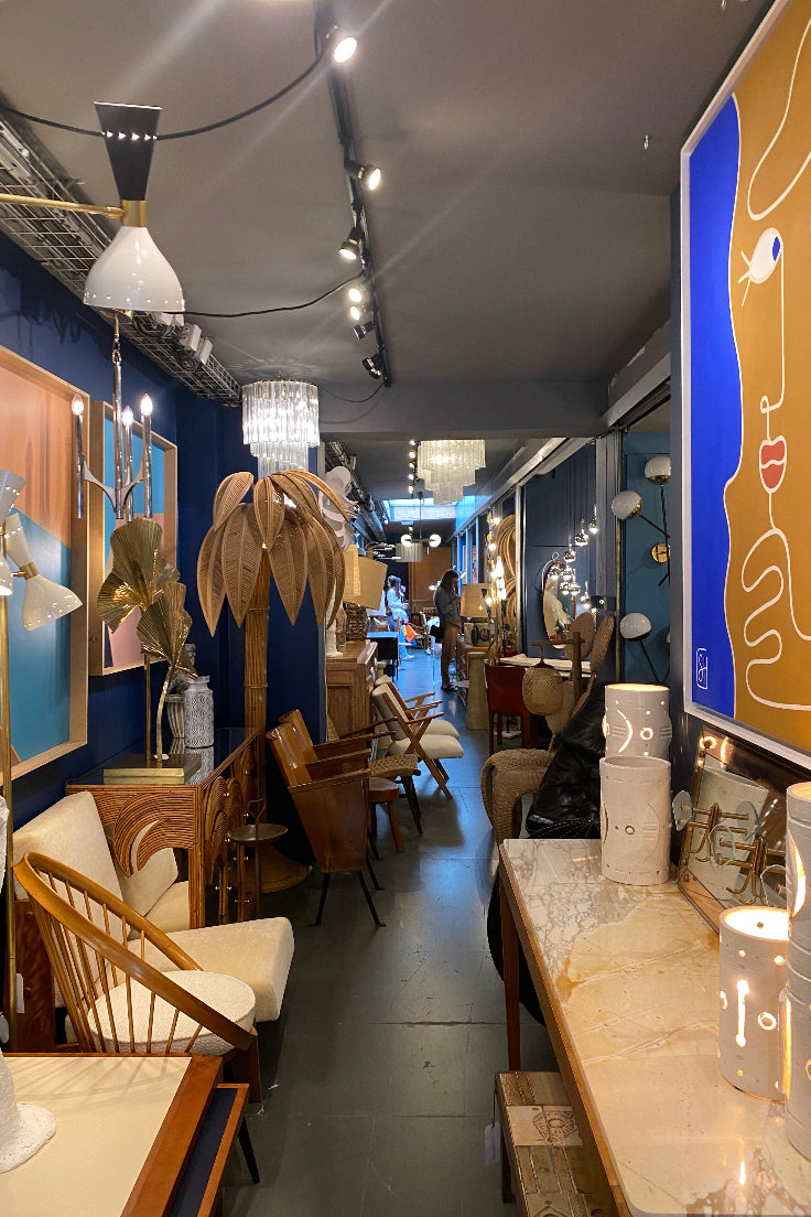A wide interior showroom scene with multiple furniture pieces and decor items arranged in a gallery-like space. Visible items include wooden chairs, tables, lamps, framed art, and sculptural decor.