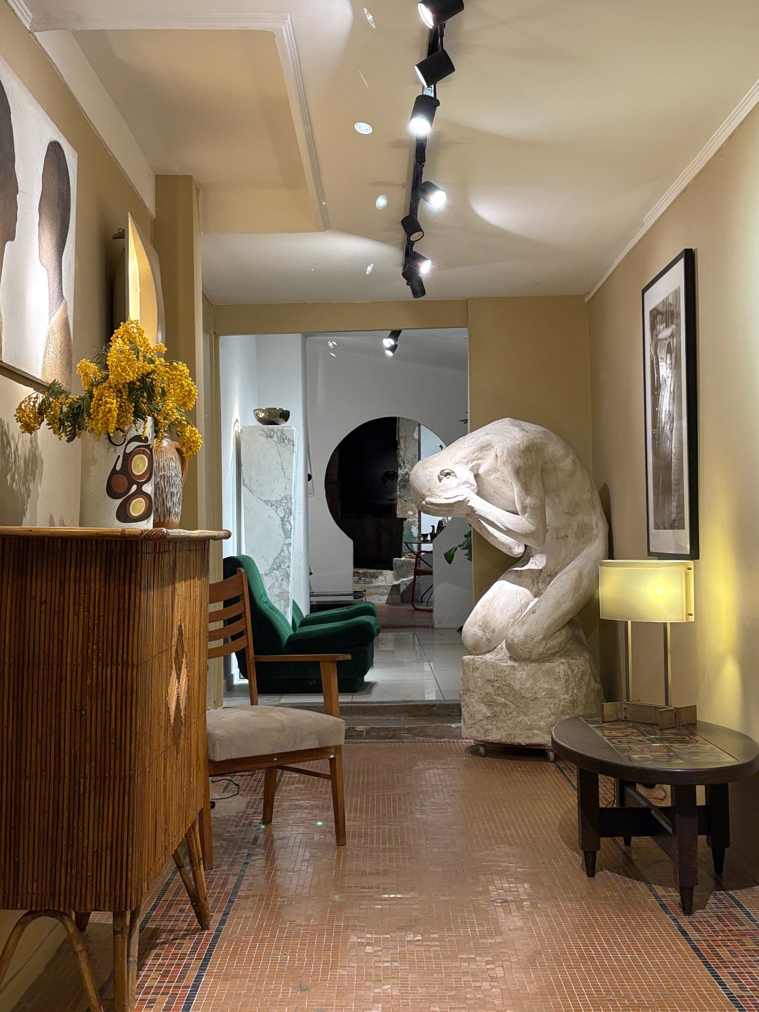 A modern dining room interior with warm lighting, featuring a wooden slatted table in the foreground, a white large-size sculpture of a man, a green velvet chair, and a brick-patterned floor.