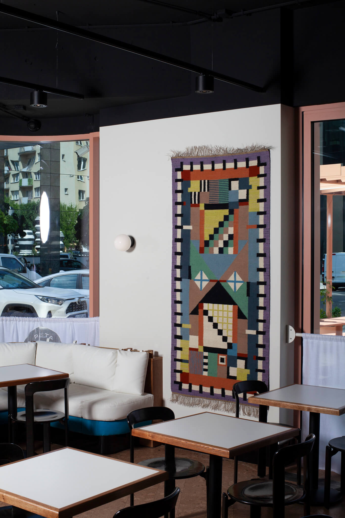 Colorful geometric-patterned rug (or kilim) on a wall in a restaurant setting with tables and chairs. Designed by Piotr Niklas for Splot via Ad Origine.